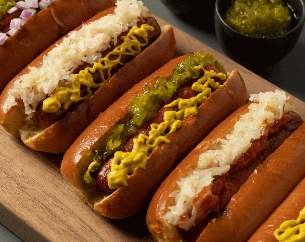 Row of hot dogs topped with mustard and relish on a wooden board