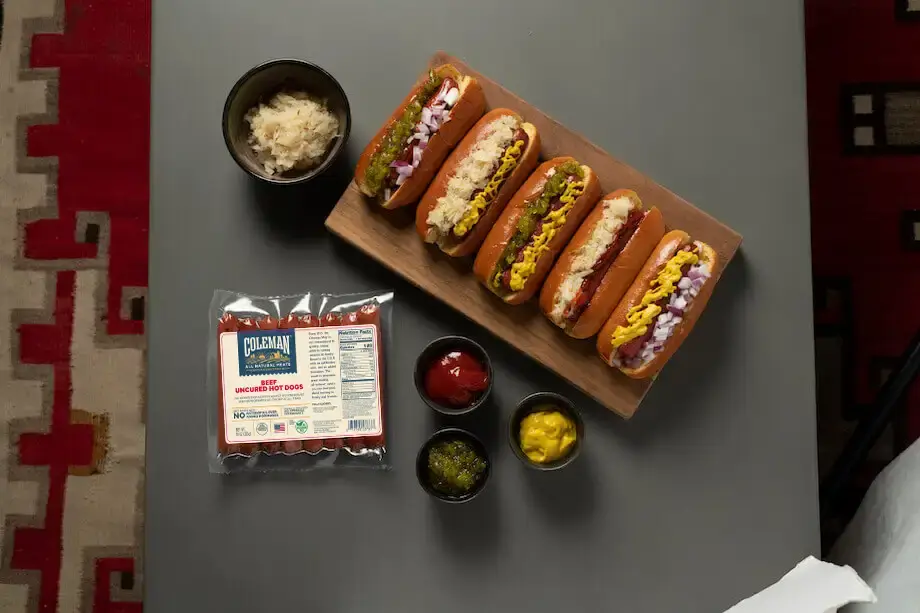 Pack of Coleman hot dogs next two five cooked hot dogs on a wooden plate